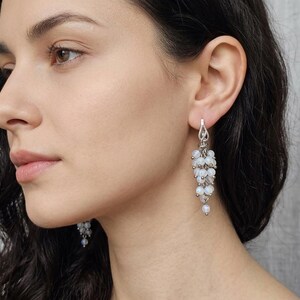 May include: A pair of silver-toned dangle earrings featuring a cluster of small, translucent, iridescent white beads arranged in a grape-like bunch. The earrings are suspended from a simple silver-toned hook and feature a delicate, multi-strand design.