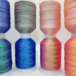 Może przedstawiać: Sześć szpulek iryzującej nici w różnych kolorach, w tym niebieskim, różowym i pomarańczowym. Nić jest nawinięta na białe plastikowe szpulki.