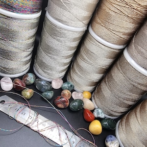May include: Six spools of metallic thread in various shades of brown, silver, and gold. The spools are arranged in a row, with the thread unwinding from each spool. A ruler and a collection of colorful stones are also visible in the image.