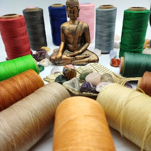 Può includere: Una statua dorata di Buddha si trova al centro di un cerchio di bobine di filo di vari colori, tra cui rosso, verde, blu, grigio e beige. Ci sono anche diverse piccole pietre colorate sparse intorno alla statua.