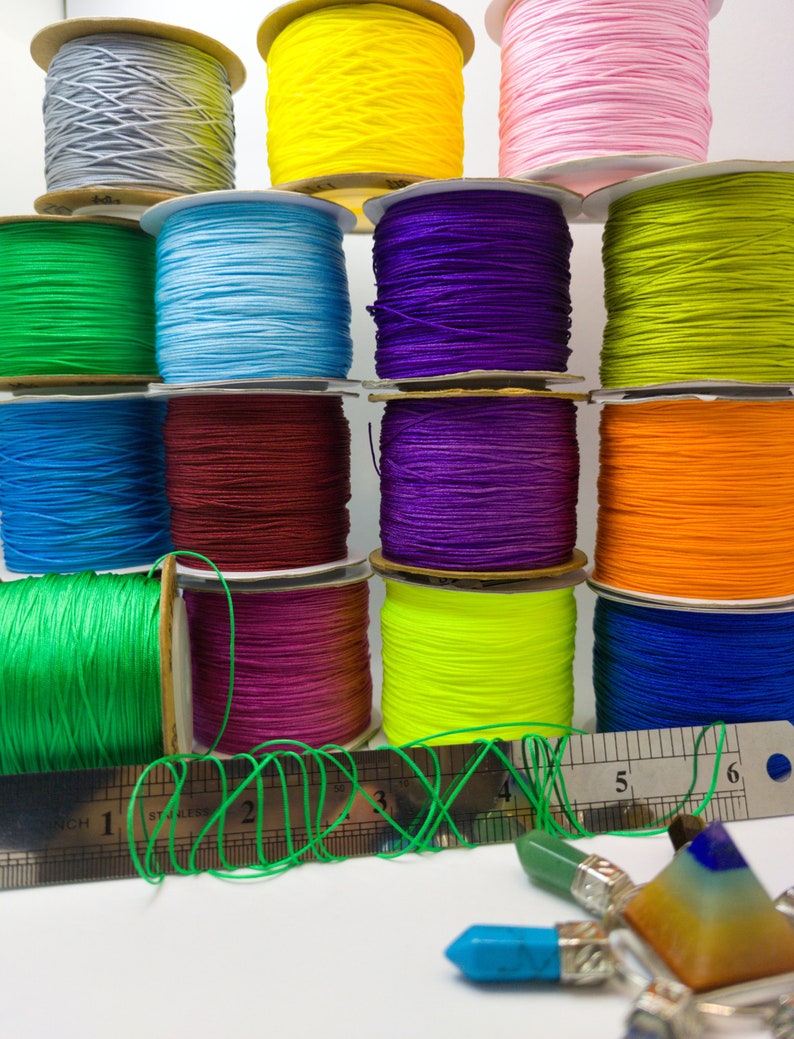 May include: Assortment of colorful spools of waxed cord, arranged in a pyramid shape. The cords are in various colors, including green, blue, purple, red, orange, yellow, and gray. A ruler is in the foreground, with a green cord wrapped around it.