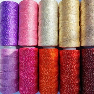 May include: Ten spools of colorful thread with a sparkly finish. The thread is wound around spools with white plastic caps. The colors are purple, pink, cream, gold, brown, lavender, hot pink, orange, red, and burgundy.