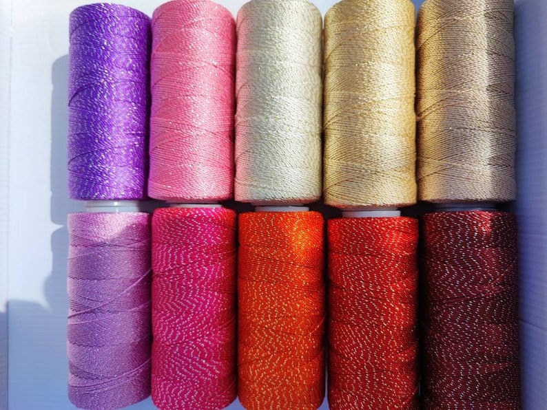 May include: Ten spools of colorful thread with a sparkly finish. The thread is wound around spools with white plastic caps. The colors are purple, pink, cream, gold, brown, lavender, hot pink, orange, red, and burgundy.