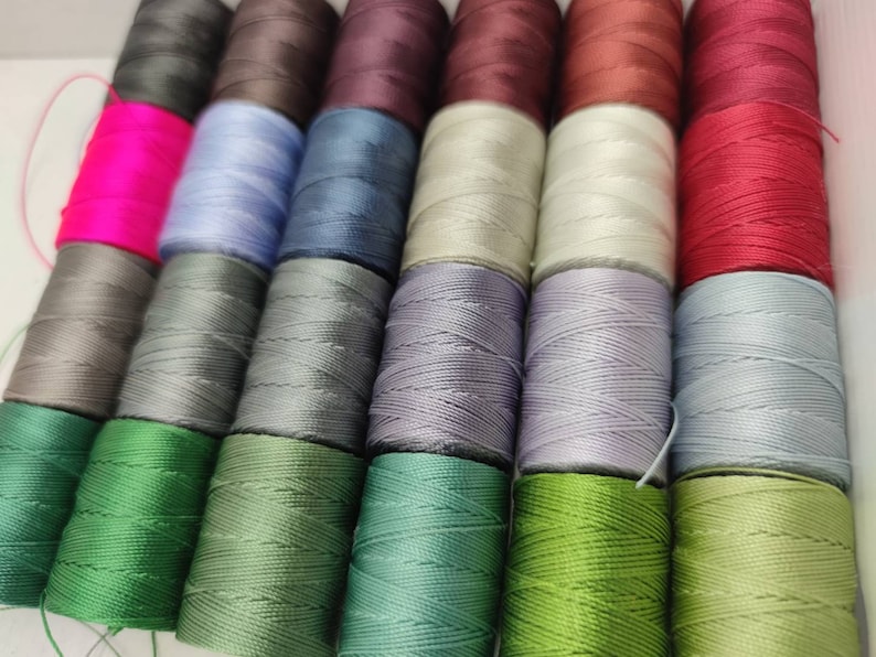 Peut inclure: Une collection de bobines de fil de diff&eacute;rentes couleurs, notamment rose, bleu, gris, vert et rouge. Les bobines sont dispos&eacute;es en rang&eacute;es et sont toutes de la m&ecirc;me taille.