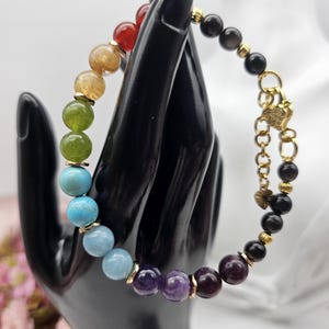 May include: A multi-colored beaded bracelet featuring a sequence of round stones in red, yellow, green, blue, light blue, and purple, transitioning into black beads. The bracelet includes gold-toned metal spacers and a gold-toned lobster clasp.