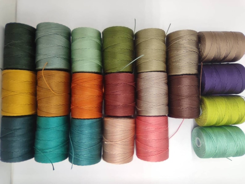 Peut inclure: Une collection de 20 bobines de fil de lin cir&eacute; de diff&eacute;rentes couleurs, notamment vert, marron, orange, rouge, beige et violet. Les bobines sont dispos&eacute;es en deux rang&eacute;es, la rang&eacute;e sup&eacute;rieure &eacute;tant l&eacute;g&egrave;rement d&eacute;cal&eacute;e par rapport &agrave; la rang&eacute;e inf&eacute;rieure.
