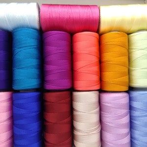 Könnte beinhalten: Eine Sammlung von 18 Garnrollen in verschiedenen Farben, darunter Rosa, Blau, Lila, Orange, Gelb und Grün. Die Rollen sind in drei Reihen zu sechs angeordnet.