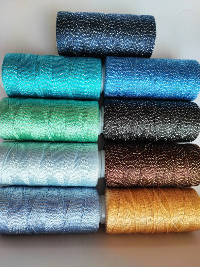 May include: A set of ten spools of thread with metallic accents. The spools are arranged in a stack, with the colors ranging from dark blue to gold.