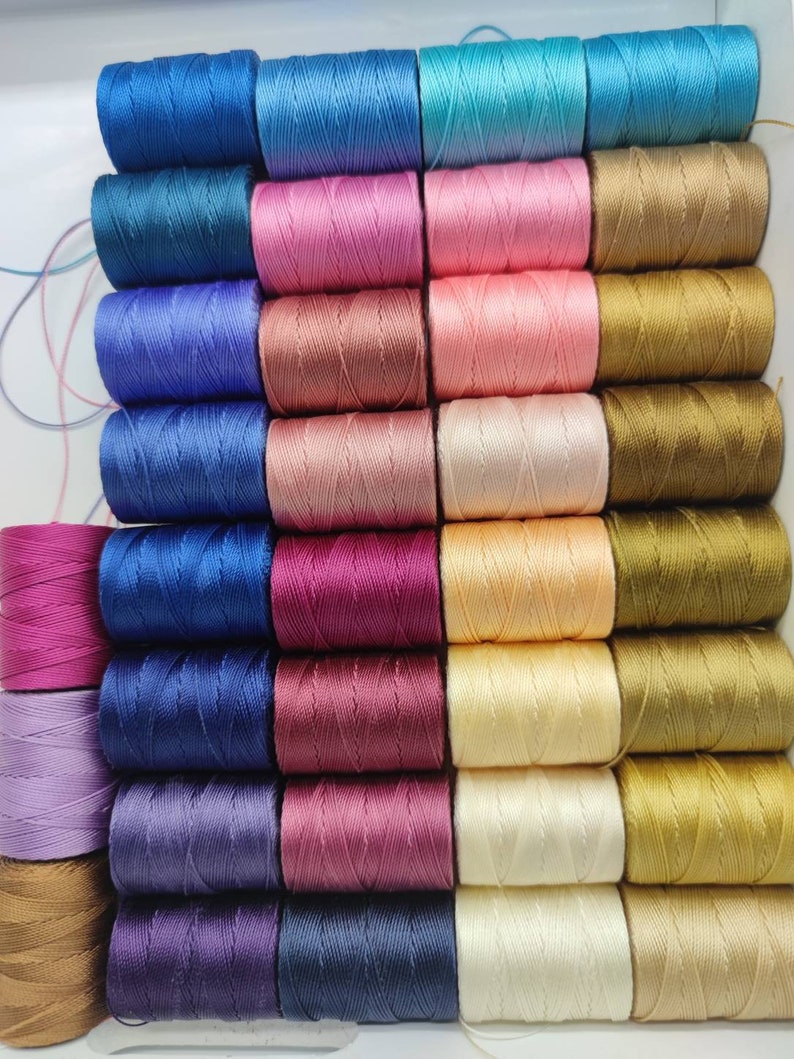 Peut inclure: Une collection de 24 bobines de fil de diff&eacute;rentes couleurs, notamment bleu, rose, marron, or, violet et rouge. Les bobines sont dispos&eacute;es en rang&eacute;es et en colonnes, et elles sont toutes de la m&ecirc;me taille.