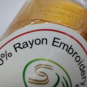 Puede incluir: Una bobina de hilo de bordar de ray&oacute;n 100% en color amarillo brillante. La etiqueta dice "100% Rayon Embroidery".