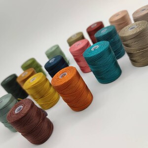 Peut inclure: Une collection de bobines de fil de diff&eacute;rentes couleurs, notamment vert, bleu, orange, marron et beige. Les bobines sont dispos&eacute;es en rang&eacute;e, les couleurs passant progressivement du vert au beige.