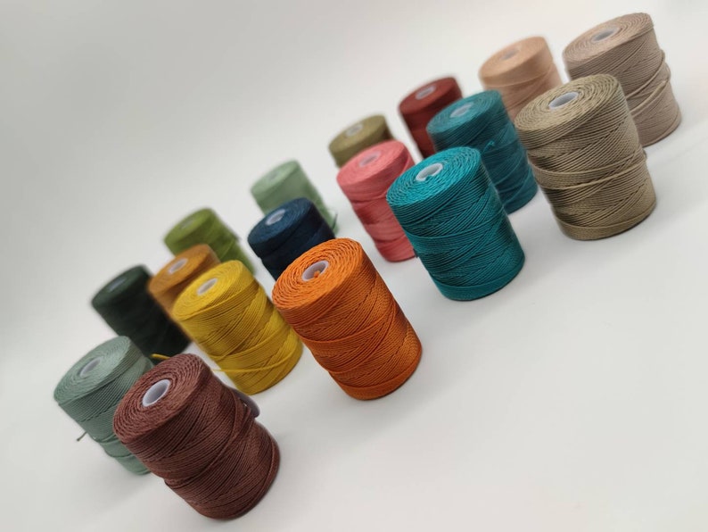 Peut inclure: Une collection de bobines de fil de diff&eacute;rentes couleurs, notamment vert, bleu, orange, marron et beige. Les bobines sont dispos&eacute;es en rang&eacute;e, les couleurs passant progressivement du vert au beige.