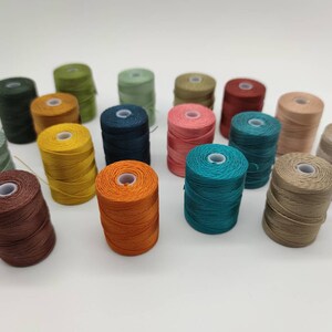 Peut inclure: Une collection de 18 bobines de fil &agrave; broder de diff&eacute;rentes couleurs, notamment turquoise, marron, orange, jaune, vert et rose. Les bobines sont dispos&eacute;es en demi-cercle sur une surface blanche.