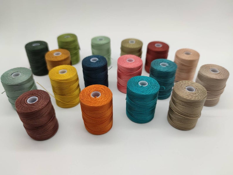 Peut inclure: Une collection de 18 bobines de fil &agrave; broder de diff&eacute;rentes couleurs, notamment turquoise, marron, orange, jaune, vert et rose. Les bobines sont dispos&eacute;es en demi-cercle sur une surface blanche.
