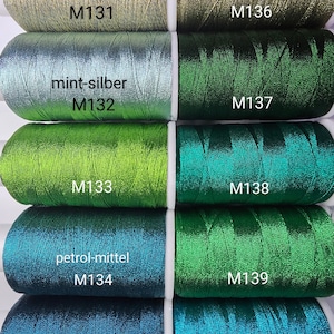 May include: A collection of spools of metallic thread in various shades of green, teal, and gold. Each spool is labeled with a number and a color description, such as "mint-silber" and "petrol-mittel". The threads have a shiny, reflective quality.