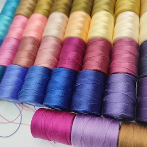 Peut inclure: Une collection de bobines de fil de diff&eacute;rentes couleurs, notamment bleu, rose, violet, rouge et marron. Les bobines sont dispos&eacute;es en rang&eacute;es et sont toutes de la m&ecirc;me taille.