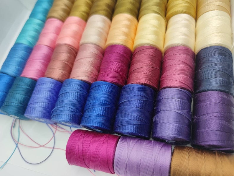 Peut inclure: Une collection de bobines de fil de diff&eacute;rentes couleurs, notamment bleu, rose, violet, rouge et marron. Les bobines sont dispos&eacute;es en rang&eacute;es et sont toutes de la m&ecirc;me taille.
