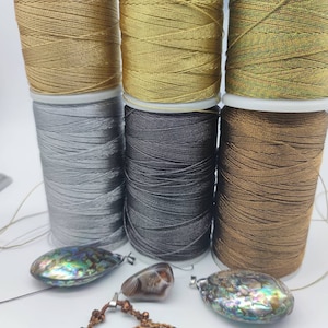 Può includere: Sei rocchetti cilindrici di filo metallico nelle tonalità oro, argento, grigio scuro e bronzo, disposti su due file. Sotto i rocchetti si trovano due ciondoli in conchiglia di abalone iridescente, una piccola pietra d'agata marrone lucida e una collana in cordino intrecciato.
