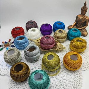 Könnte beinhalten: Sortiment an bunten Garnknäueln in verschiedenen Farbtönen von Blau, Grün, Rot, Gold, Silber, Weiß, Lila und Braun. Die Garnknäuel sind auf einer weißen Häkeldeckchen angeordnet.