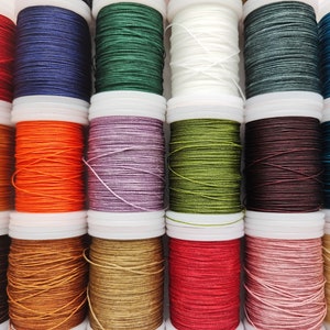 Könnte beinhalten: Eine Sammlung von 12 Garnrollen in verschiedenen Farben, darunter Rot, Orange, Gelb, Grün, Blau, Lila, Braun und Weiß. Die Rollen sind in einer Reihe angeordnet, wobei das Garn um jede Rolle gewickelt ist.