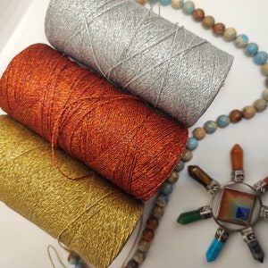 Könnte beinhalten: Drei zylindrische Garnrollen aus Metallic-Garn in Silber, Kupfer und Gold, die diagonal angeordnet sind. Eine Schnur aus mehrfarbigen Steinperlen verläuft neben den Garnrollen und endet bei einem sternförmigen Anhänger mit sieben Kristallspitzen.