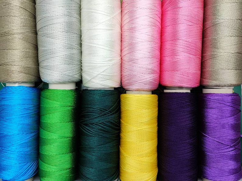 Puede incluir: Una colecci&oacute;n de carretes de hilo en varios colores, incluyendo beige, gris, blanco, rosa, azul, verde, verde oscuro, amarillo, morado y lavanda.
