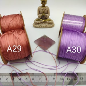 May include: Two spools of cord, one pink and one purple, with the labels A29 and A30. The spools are sitting on a white surface with a small golden statue of Buddha in the center.