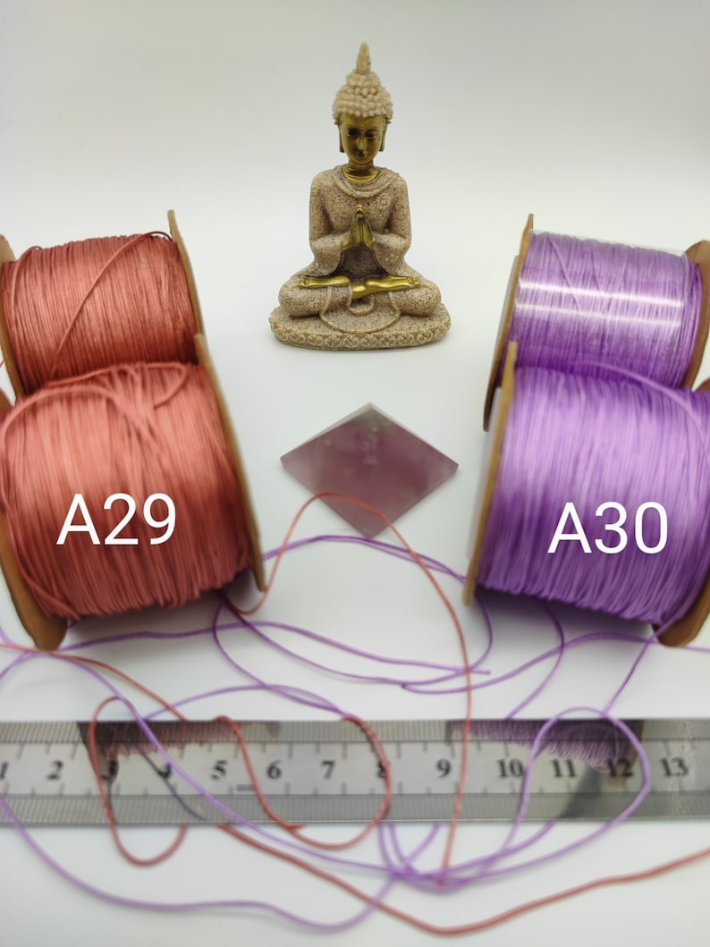 May include: Two spools of cord, one pink and one purple, with the labels A29 and A30. The spools are sitting on a white surface with a small golden statue of Buddha in the center.