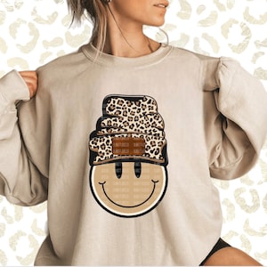 May include: A beige sweatshirt with a smiling face graphic wearing a brown and black leopard print beanie.