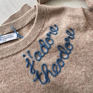May include: A beige long-sleeve sweater featuring blue embroidered cursive text that reads "j'adore theodore". The garment has a ribbed crew neckline and a white interior tag labeled "monica threads" and "ty cashmere" with the size "small".