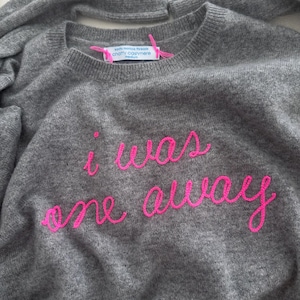 May include: A gray crew neck sweater with the embroidered phrase "i was one away" in bright pink cursive lettering. The sweater has a tag that reads "santa monica threads chatty cashmere medium".