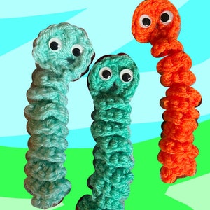 Puede incluir: Tres juguetes de gusano de crochet con ojos saltones. Un gusano es naranja y los otros dos son de color turquesa.