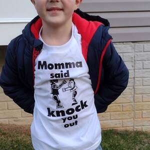 May include: A young boy wearing a white t-shirt with a black graphic that says "Momma said knock you out". He is also wearing a blue jacket with a red hood, blue jeans, and brown boots.