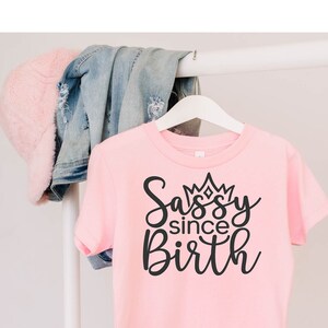 May include: Pink t-shirt with black text that reads "Sassy since Birth" with a crown above the word "Sassy".
