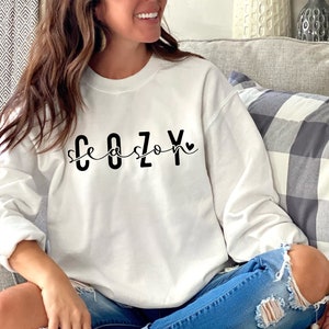 Puede incluir: Una sudadera blanca con el texto "Cozy Season" en letras negras con un peque&ntilde;o coraz&oacute;n al final del texto.