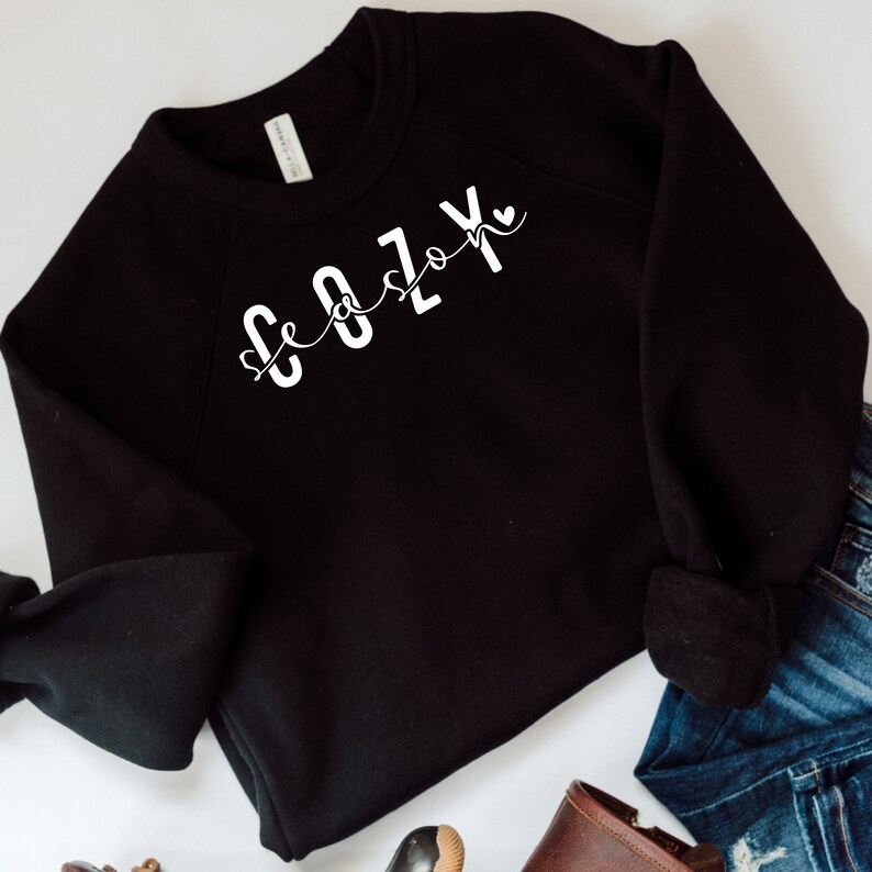 Puede incluir: Sudadera negra con texto blanco que dice "Cozy Season" con un peque&ntilde;o coraz&oacute;n al final del texto.
