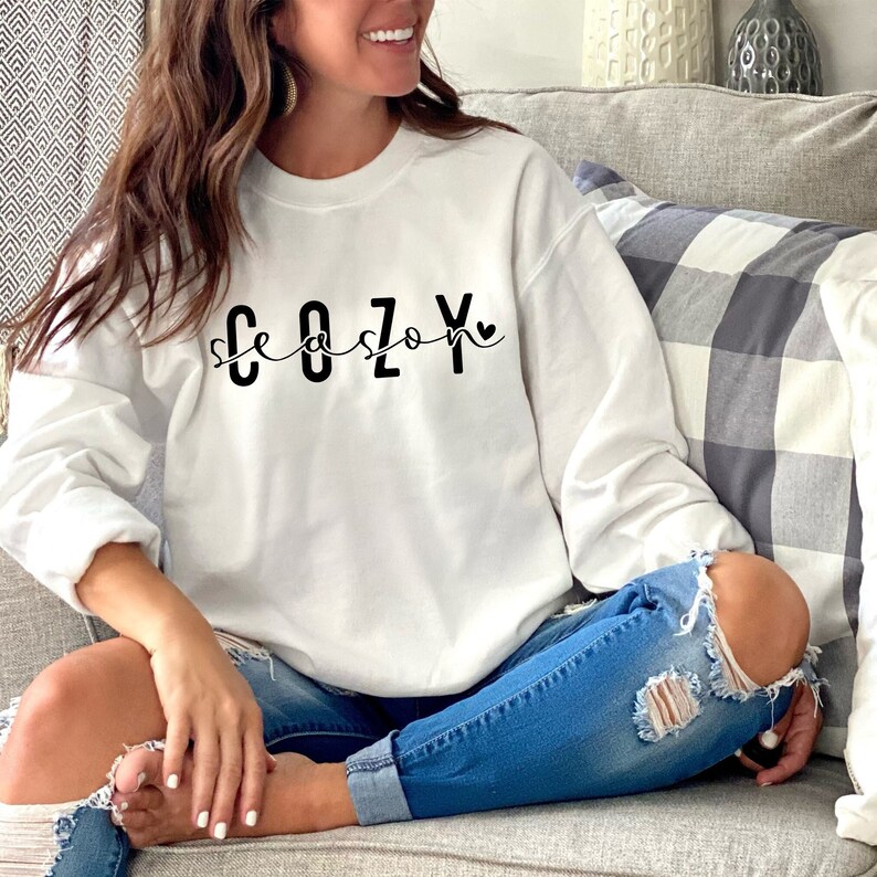 Puede incluir: Una sudadera blanca con el texto "Cozy Season" en letras negras con un peque&ntilde;o coraz&oacute;n al final del texto.