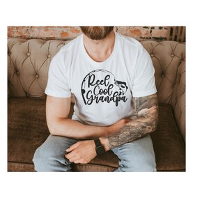 K&ouml;nnte beinhalten: Wei&szlig;es T-Shirt mit dem Text "Reel Cool Grandpa" in schwarzer, stilisierter Schrift. Das Design beinhaltet eine Angelrute und einen Fisch. Das Shirt ist kurz&auml;rmlig.