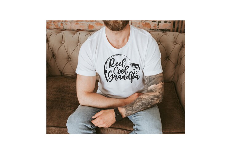 K&ouml;nnte beinhalten: Wei&szlig;es T-Shirt mit dem Text "Reel Cool Grandpa" in schwarzer, stilisierter Schrift. Das Design beinhaltet eine Angelrute und einen Fisch. Das Shirt ist kurz&auml;rmlig.