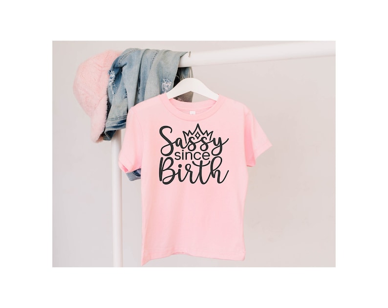 May include: Pink t-shirt with black text that reads "Sassy since Birth" with a crown above the word "Sassy".