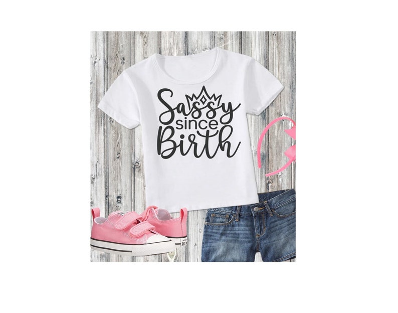 May include: White t-shirt with black text that says "Sassy since Birth" with a crown above the word "Sassy". The shirt is on a wooden background with pink shoes and denim shorts.