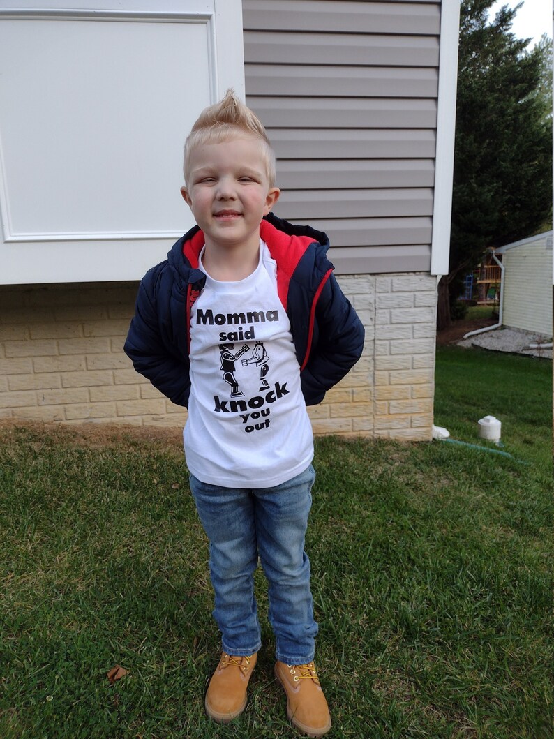 May include: A young boy wearing a white t-shirt with a black graphic that says "Momma said knock you out". He is also wearing a blue jacket with a red hood, blue jeans, and brown boots.