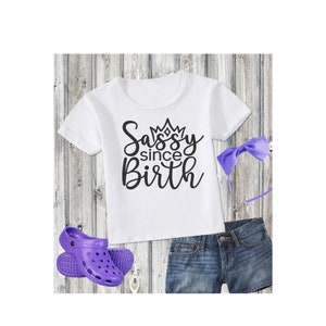 May include: White t-shirt with black text that says "Sassy since Birth" with a crown above the word "Sassy". The shirt is paired with purple denim shorts, a purple headband, and purple clogs.