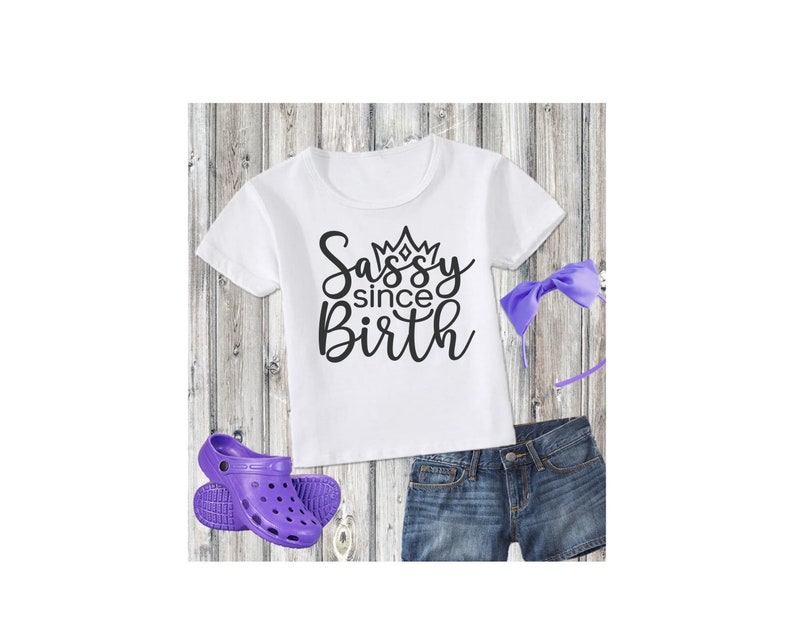 May include: White t-shirt with black text that says "Sassy since Birth" with a crown above the word "Sassy". The shirt is paired with purple denim shorts, a purple headband, and purple clogs.