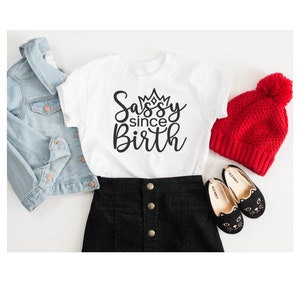 May include: White t-shirt with a black graphic that says "Sassy since Birth" with a crown above the word "Sassy". The shirt is paired with a black skirt, a denim jacket, a red beanie, and black cat-shaped shoes.