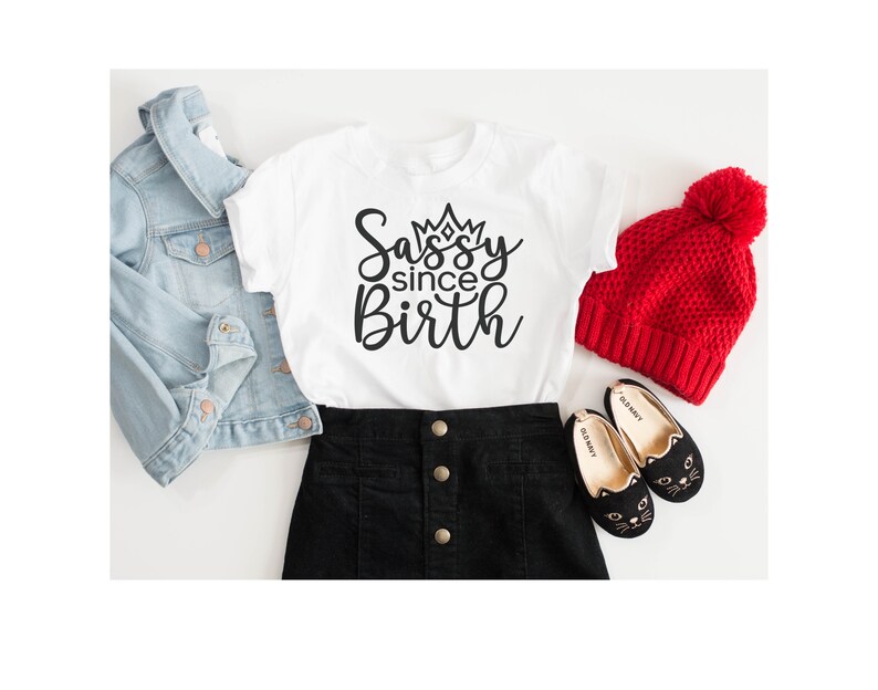 May include: White t-shirt with a black graphic that says "Sassy since Birth" with a crown above the word "Sassy". The shirt is paired with a black skirt, a denim jacket, a red beanie, and black cat-shaped shoes.