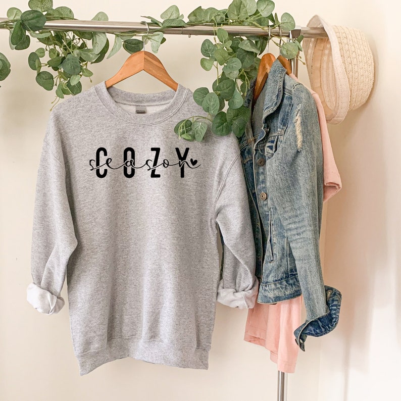 Puede incluir: Una sudadera gris con el texto "Cozy Season" en letras negras y un peque&ntilde;o s&iacute;mbolo de coraz&oacute;n.
