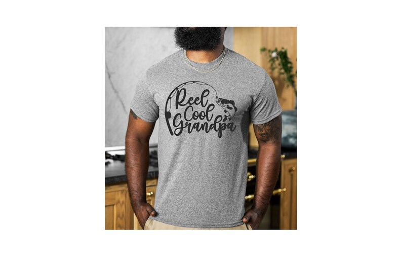K&ouml;nnte beinhalten: Ein graues T-Shirt mit dem Text "Reel Cool Grandpa" in schwarzer Schrift und einer Angelruten-Grafik. Das kurz&auml;rmelige Shirt ist aus weichem Material. Das Design beinhaltet einen Fisch und eine Angelrute.