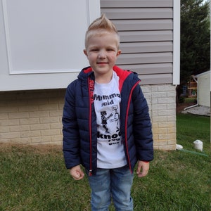 May include: A young boy wearing a white t-shirt with a black and white graphic of two robots boxing. The shirt says "Momma said knock you out". He is also wearing a navy blue puffer jacket with red zipper pulls and blue jeans.