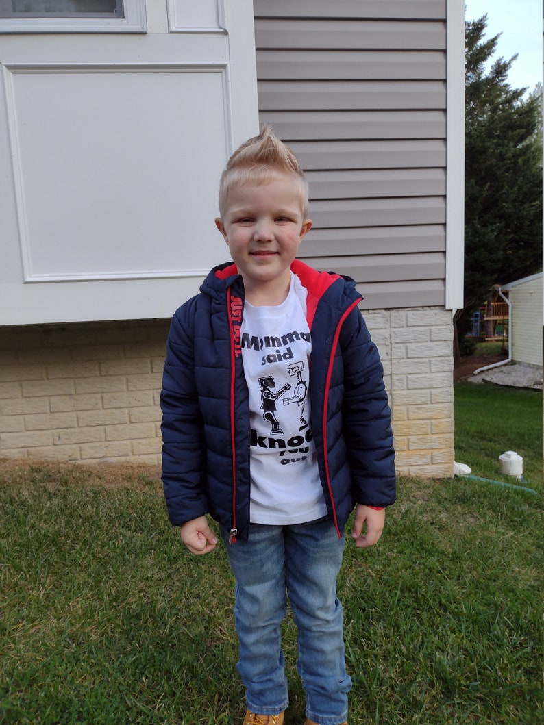 May include: A young boy wearing a white t-shirt with a black and white graphic of two robots boxing. The shirt says "Momma said knock you out". He is also wearing a navy blue puffer jacket with red zipper pulls and blue jeans.